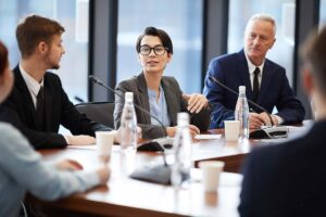 Uma jovem empresária falando em uma reunião enquanto discute projetos de governança corporativa e compliance com quatro colegas sentados à mesa redonda,.