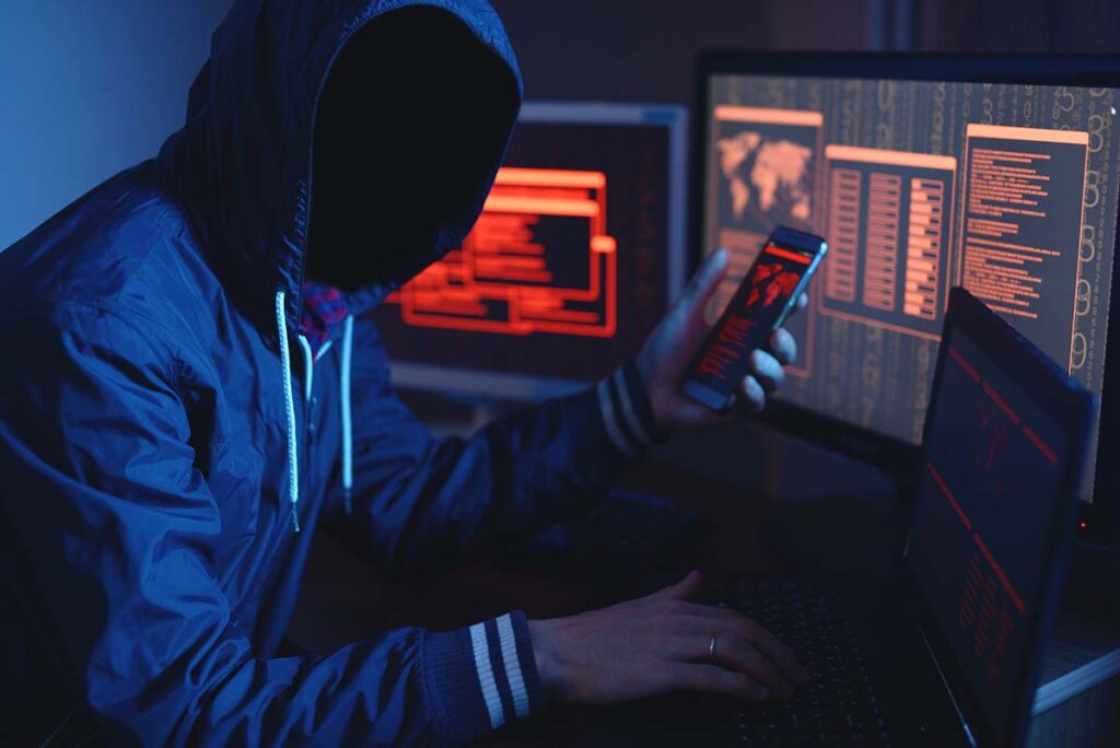A imagem mostra um homem com o rosto encoberto por um capuz, como se fosse um criminoso, mexendo em dispositivos eletrônicos (computador, notebook e smartphone).
