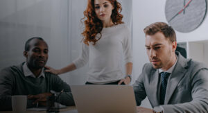 Uma mulher está em pé enquanto dois homens estão sentados, um digitando no teclado de um notebook e o outro observando seu trabalho, trazendo o contexto de análise de risco nas empresas.