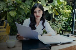 Mulher segura papéis enquanto está sentada à frente de uma mesa com outros documentos, pastas e celular sobre a mesa.