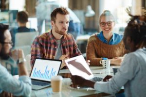 Um grupo de quatro profissionais conversam em um escritório enquanto estão sentados em cadeiras corporativas, trazendo o contexto da integração entre os times de marketing e vendas.