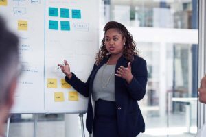 Mulher negra está em pé, com um quadro branco repleto de dados, explicando para pessoas que estão sentadas em uma sala de reunião corporativa, trazendo o contexto da estratégia de expansão de mercado.