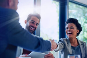 Um homem sentado sorri enquanto segura um papel, um outro homem em pé cumprimenta uma mulher sentada, todos em um ambiente corporativo, trazendo a ideia de que vão participar de uma reunião sobre ciclo de vendas B2B.
