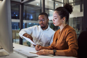 Dupla de trabalho, sendo um homem negro e uma mulher parda, trabalham juntos em um documento à frente de um computador, contextualizando o tema de verificação KYC, tema deste artigo.