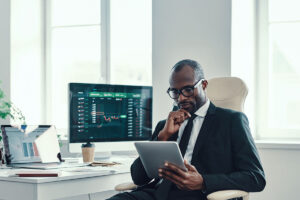 Homem negro segurando um tablet em enquanto está sentado em uma cadeira, aparentemente em seu trabalho, com uma tela de computador repleta de gráficos atrás - os dados do gráficos podem ter relação com gestão de investimentos.