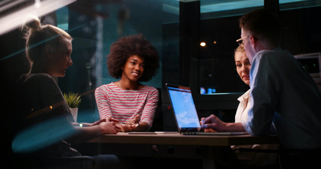 Foto de quatro pessoas ao redor de uma mesa de reuniões, em um escritório e com um laptop, ilustra este artigo sobre como a tecnologia aprimora o processo de diligências nas empresas.