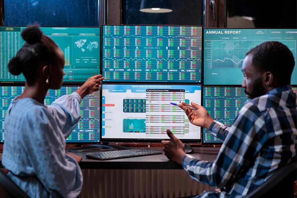 Mulher e homem conversam sobre informações exibidas em monitores, em fotografia que ilustra este artigo sobre mercado de capitais.