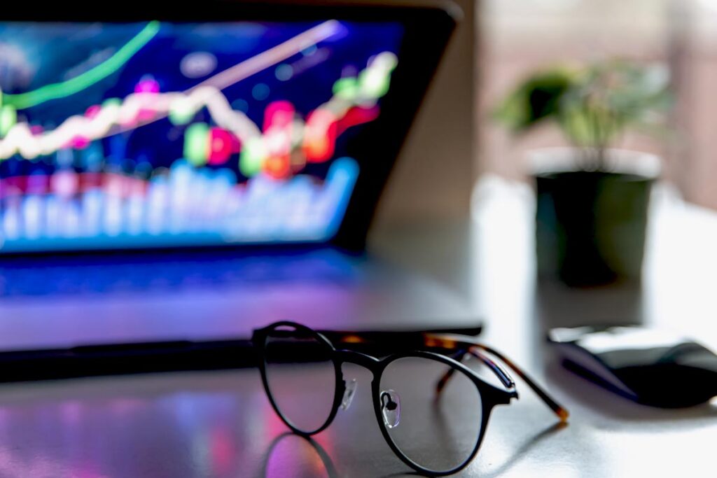 Óculos de grau sobre uma mesa. Ao fundo, a tela de um laptop exibe gráficos, em fotografia que ilustra este artigo sobre mercado de capitais.
