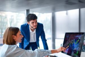 Homem e mulher observando um monitor com gráficos, enquanto a mulher aponta para a tela, provavelmente querendo entender como as empresas devem avaliar os riscos dos investimentos.