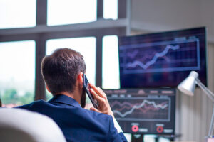 Homem sentado fala ao celular, enquanto monitores de computador mostram gráficos em suas telas para ilustrar este artigo sobre perfil de investidor.