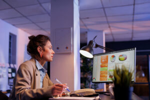 Mulher branca está com uma caneta nas mãos enquanto observa o monitor de seu computador, supostamente com dados de projeção de vendas.