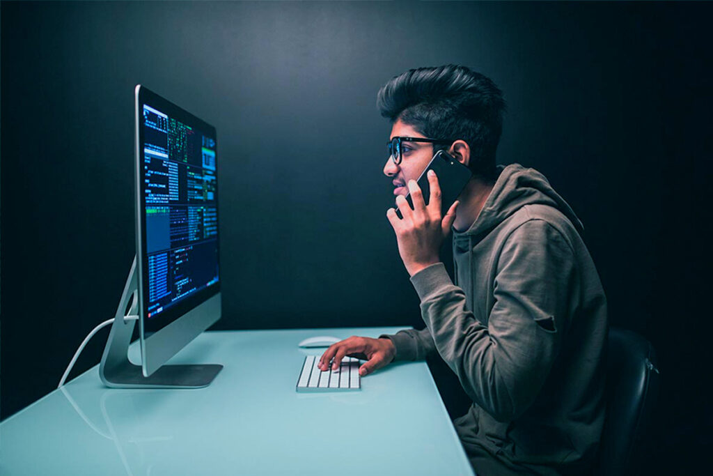 Homem jovem, de óculos, fala em um celular e mexe em um computador com a outra mão enquanto olha para uma tela.