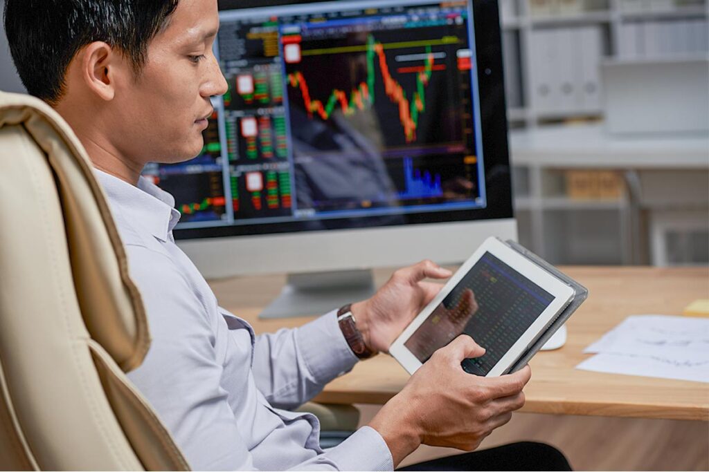 Homem sentado mexe em tablet, enquanto monitores de computador mostram gráficos em suas telas para ilustrar este artigo sobre perfil de investidor.