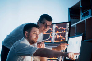 Dois homens apontam para um monitor, em fotografia que ilustra este artigo sobre como entender e acompanhar o mercado de balcão.