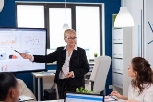 Mulher aponta para um monitor com gráficos em uma sala de reunião, observada por pelo menos outras cinco pessoas, em fotografia que ilustra este artigo sobre como gerar novos negócios com modelagem de dados do mercado financeiro.