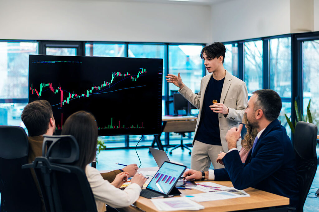 Um homem em pé apresenta análises do mercado de bolsa por instrumentos financeiros presentes em um monitor para dois homens e duas mulheres sentados em uma mesa de escritório.