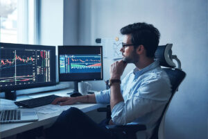 Homem observa gráficos em dois monitores, com o objetivo de entender mais sobre captura de dados no mercado de capitais.