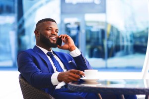Homem negro sentado segurando uma xícara de café em uma mão enquanto sorri ao conversar pelo celular com a outra mão.