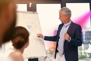 Homem branco em pé escreve em um quadro com folhas de papel enquanto um outro homem e uma mulher o observam, trazendo o contexto da importância da estratégia competitiva nas empresas. Ao fundo, uma janela com vista para a cidade.