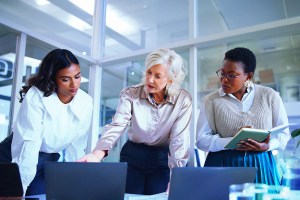 Duas mulheres negras e uma branca estão juntas em uma sala corporativa, enquanto a mulher branca aponta para a tela de um notebook e as demais observam a tela também, trazendo o contexto de discussão sobre a marcação a mercado.