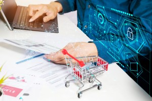 Mão de um homem digita no teclado de um notebook enquanto a outra, a qual está apoiada sobre uma mesa com outros papéis e um carrinho de compras de brinquedo, segura um papel. Há alguns dados projetados sobre a roupa do homem, trazendo o contexto de one stop shop do Neoway Seeker.