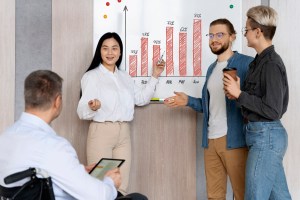 Uma mulher aponta para um quadro branco com uma caneta enquanto uma outra mulher em pé a observa ao lado de um homem também em pé e outro homem sentado observando a explicação, trazendo o contexto das estratégias de Sales Enablement nas empresas B2B.