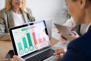 Três mulheres estão sentadas em frente a uma mesa de escritório corporativo, sendo que duas digitam em um notebook que apresenta dados de análise de vendas na tela, e a outra digita em um tablet.
