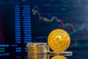 Algumas moedas douradas empilhadas com um B no centro e uma em destaque em pé sobre uma outra, com um fundo mostrando uma tela com gráficos, trazendo o contexto do Futuro de Bitcoin.