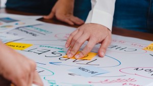Algumas mãos sobre uma mesa com cartolina e post-it, os quais possuem dados e ideias para montar um futuro planejamento comercial.