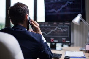 Um homem fala ao celular enquanto está sentado olhando para o monitor do computador à sua frente, o qual apresenta gráficos com dados de investimentos, trazendo contexto da importância do Raio-X do Investidor Brasileiro para instituições financeiras.