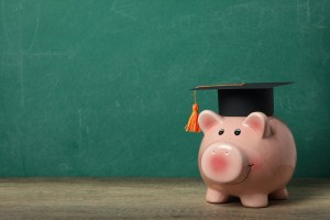 Cofre em formato de porquinho com um chapéu de formatura, capelo, com uma lousa ao fundo, trazendo o contexto de educação financeira no Brasil.