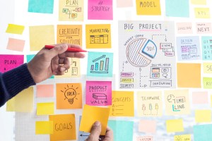Uma mão segura uma caneta enquanto a outra segura um post it, enquanto há diversos outros post it colados em uma parede de vidro, trazendo o contexto de planejamento estratégico de uma empresa B2B.