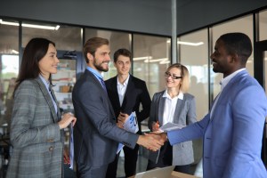Um homem branco acompanhado de outro homem branco e duas mulheres brancas, todos sorrindo em um escritório corporativo, cumprimenta um homem negro, trazendo o contexto de clientes enterprise.