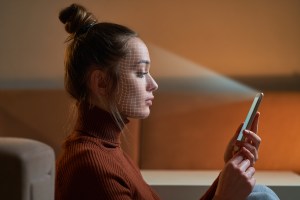 Mulher branca sentada enquanto segura com uma das mãos um celular, o qual está apontado para seu rosto como se estivesse fazendo reconhecimento facial, trazendo o contexto da tecnologia de liveness (prova de vida).