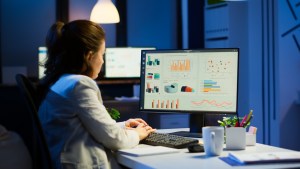 Mulher sentada digita no teclado de um computador enquanto observa o monitor, o qual apresenta diversos gráficos, trazendo o contexto de Marketing Mix Modeling nas empresas.