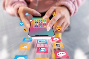 As mãos de uma mulher clicando na tela de um celular, o qual está apresentando logos de aplicativos na sua superfície, além de outros logos e emojis à frente e ao redor do aparelho, trazendo o contexto de segmentação de audiência.