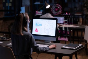 Uma mulher sentada e usando o mouse enquanto observa o monitor de um computador, o qual apresenta diversos dados e gráficos, com outros dados e gráficos holográficos ao redor da tela, trazendo o contexto de um sistema de inteligência de marketing.