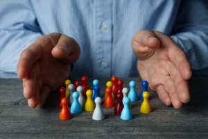 Mãos de um homem estão abertas com as palmas apoiadas em uma mesa, a qual possui pinos coloridos que representam a diversidade de pessoas, trazendo o contexto de segmentação comportamental.